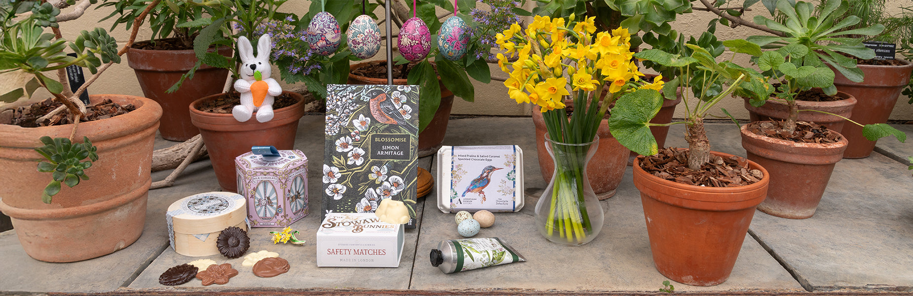 Collection of Easter themed gifts, knitted white bunny rabbit decoration, chocolate boxes, tin easter eggs and floral hand cream and books. Set against backdrop of botanical gardens pots of plants and daffodils.