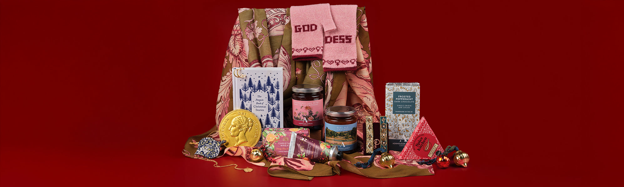 Selection of Ashmolean gifts, including mittens, scarf, books, chocolate, jams and soap against a red background