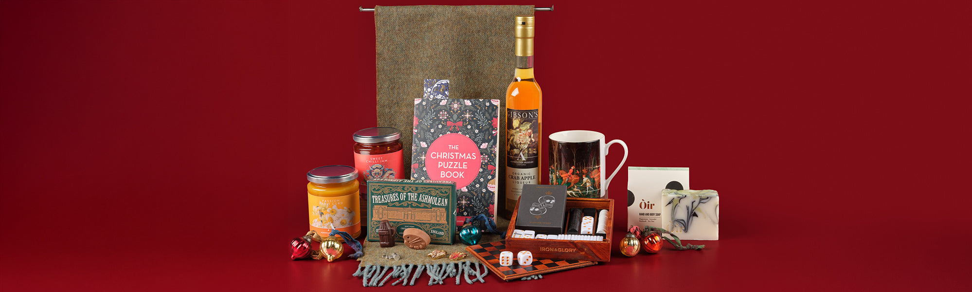 Selection of Christmas gifts, including a wool scarf, chocolates, alcoholic liqueur, soap and jams against a red background