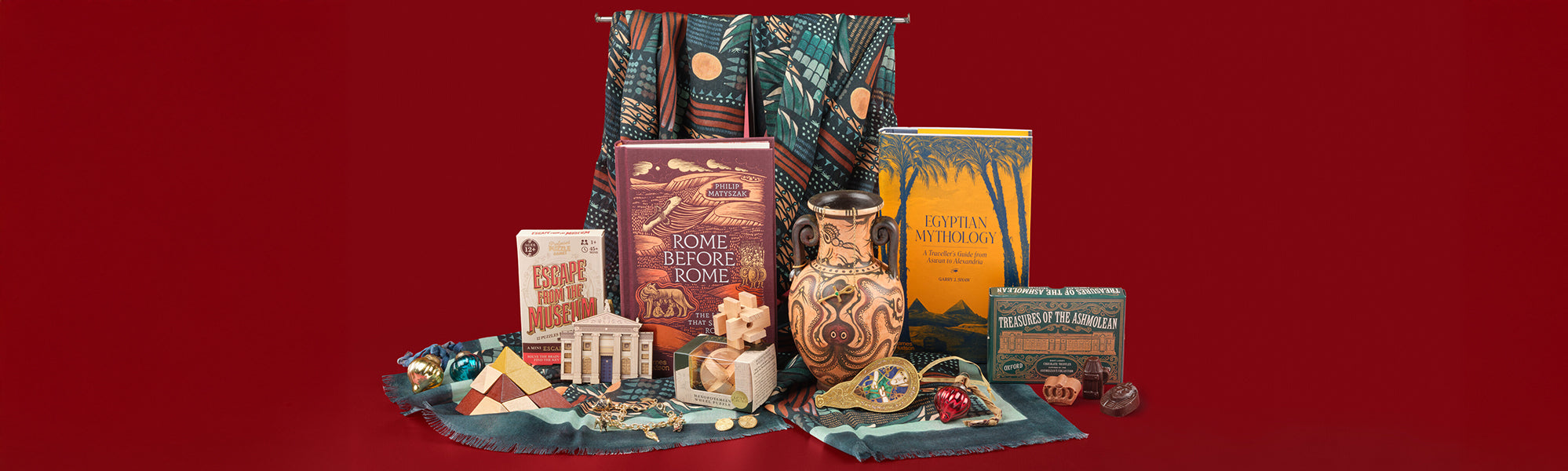 A selection of History themed gifts, including books, a vase, chocolates, decorations, puzzles and jewellery against a red background.