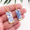 Two blue and white floral ceramic pendants on gold chains held in a hand against a white background.
