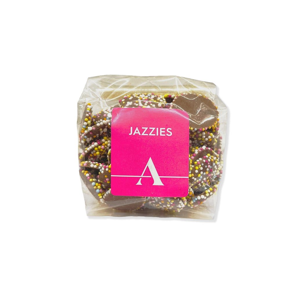A clear plastic bag containing chocolate-covered pretzels sprinkled with multicolored candy, with a bright pink square label reading 'JAZZIES' and a white stylized 'A' logo.