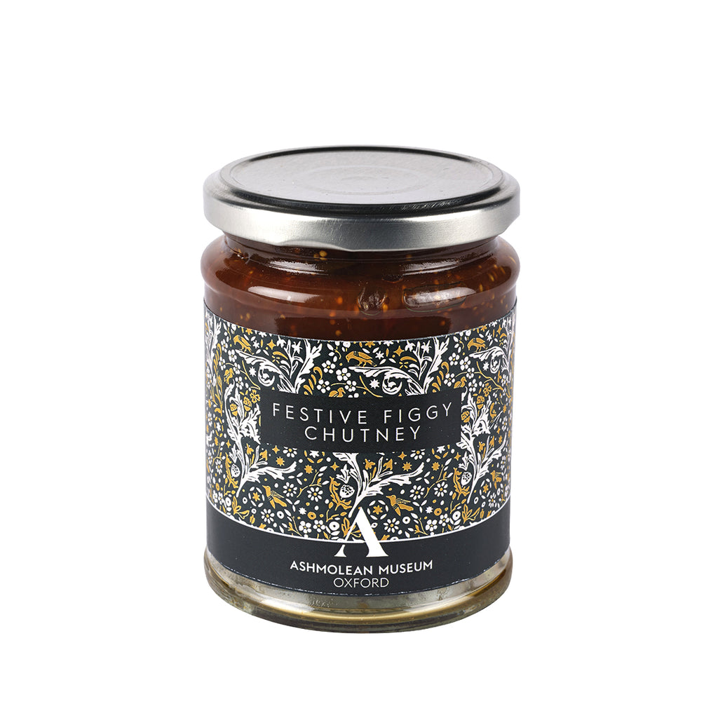 A glass jar of Festive Figgy Chutney with a silver metal lid and a black label decorated with white and gold tree illustrations, set against a plain white background.