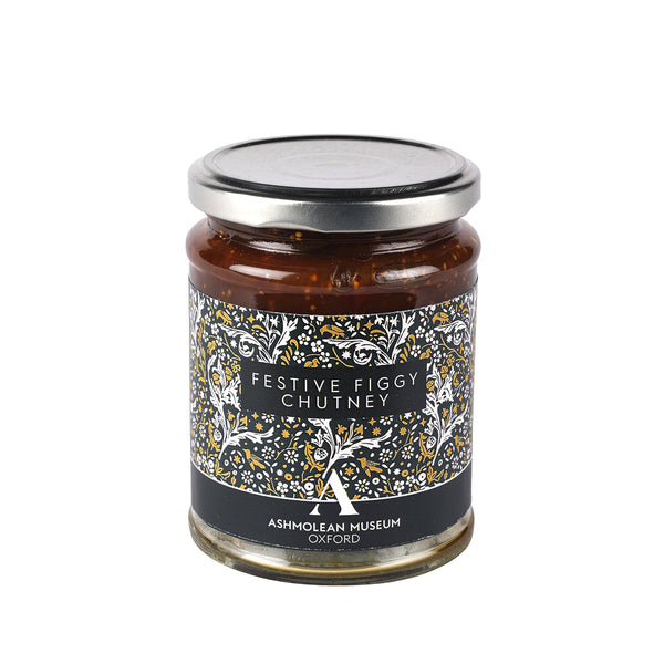 A glass jar of Festive Figgy Chutney with a silver metal lid and a black label decorated with white and gold tree illustrations, set against a plain white background.