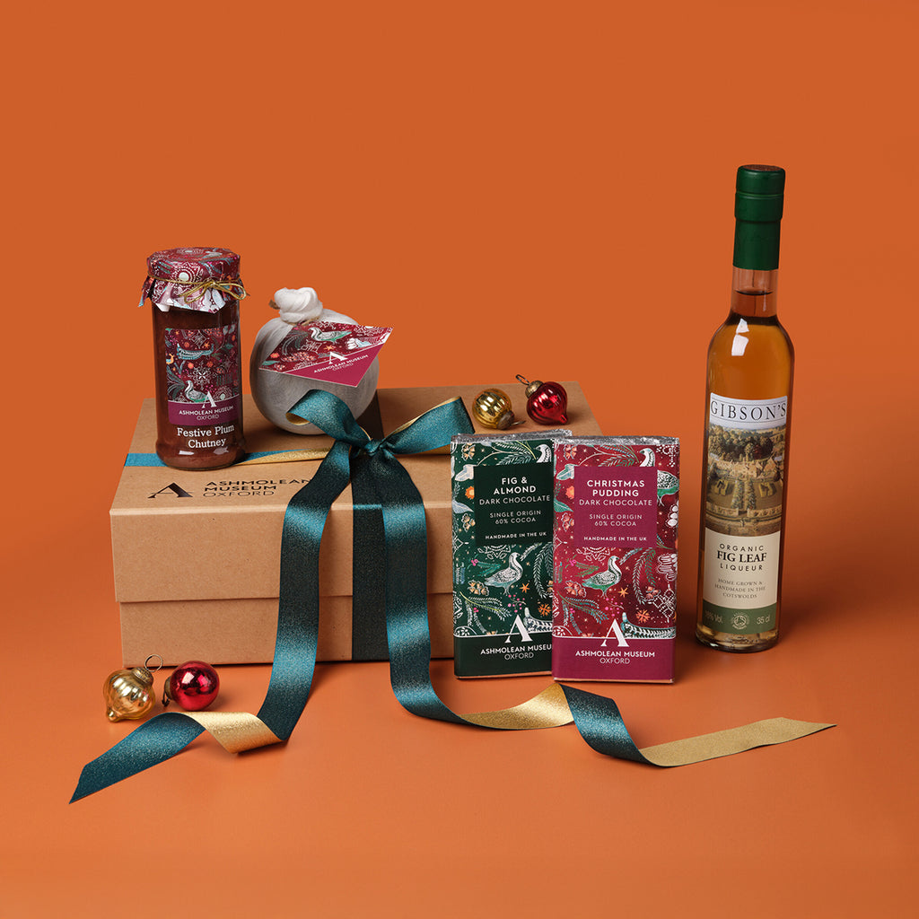 Gift box with festive items including a bottle of liqueur, jam jar, Christmas pudding, chocolate bars, and hamper box against an orange background