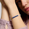 A close-up of a woman's wrist wearing a beaded bracelet with blue and white beads, a gold leaf charm, and a pearl accent, paired with a mauve dress.