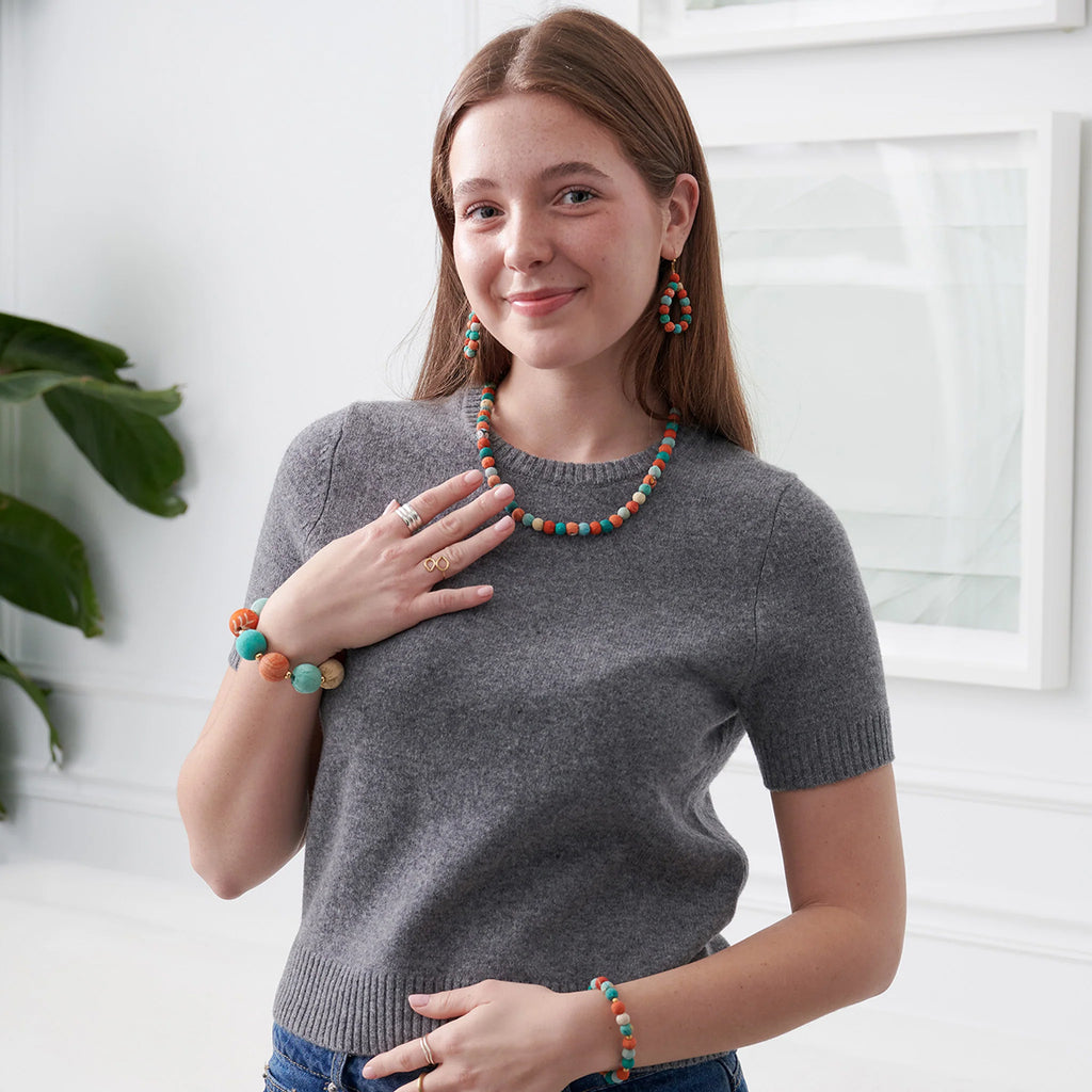 Woman wearing a gray sweater with colorful jewelry against a white wall.