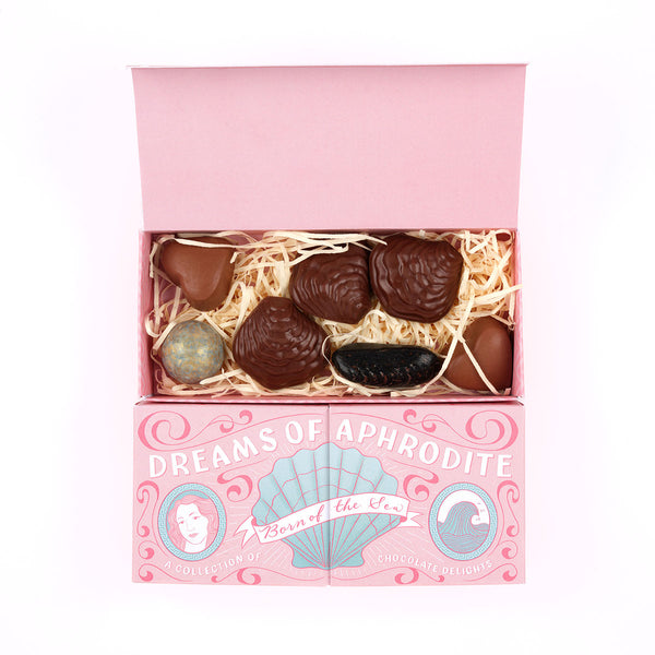 An open pink gift box filled with assorted chocolates on a white background. The box lid is pink and printed with the text 'DREAMS OF APHRODITE' and decorative shell artwork.