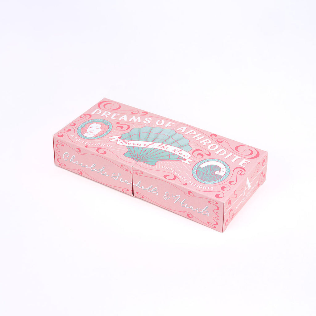 A pink decorative chocolate box with illustrated shell and swirl designs, featuring the text 'Dreams of Aphrodite' and 'Chocolate Shells & Hearts,' photographed on a clean white background.