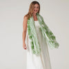 Female model wearing lightweight large green patterned scarf over all white outfit on a white background. 