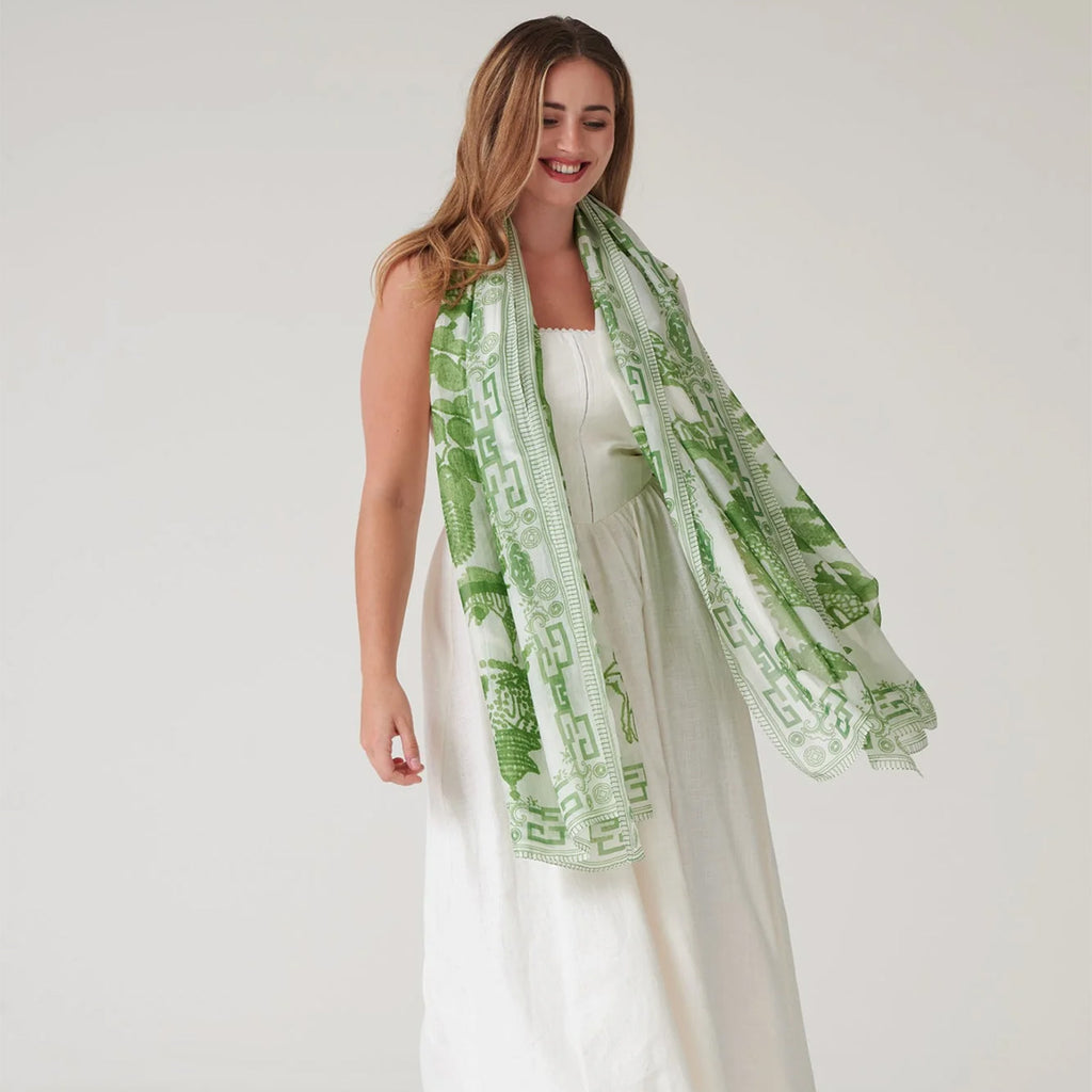 Female model wearing lightweight large green patterned scarf over all white outfit on a white background. 