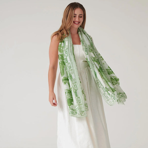Female model wearing lightweight large green patterned scarf over all white outfit on a white background. 