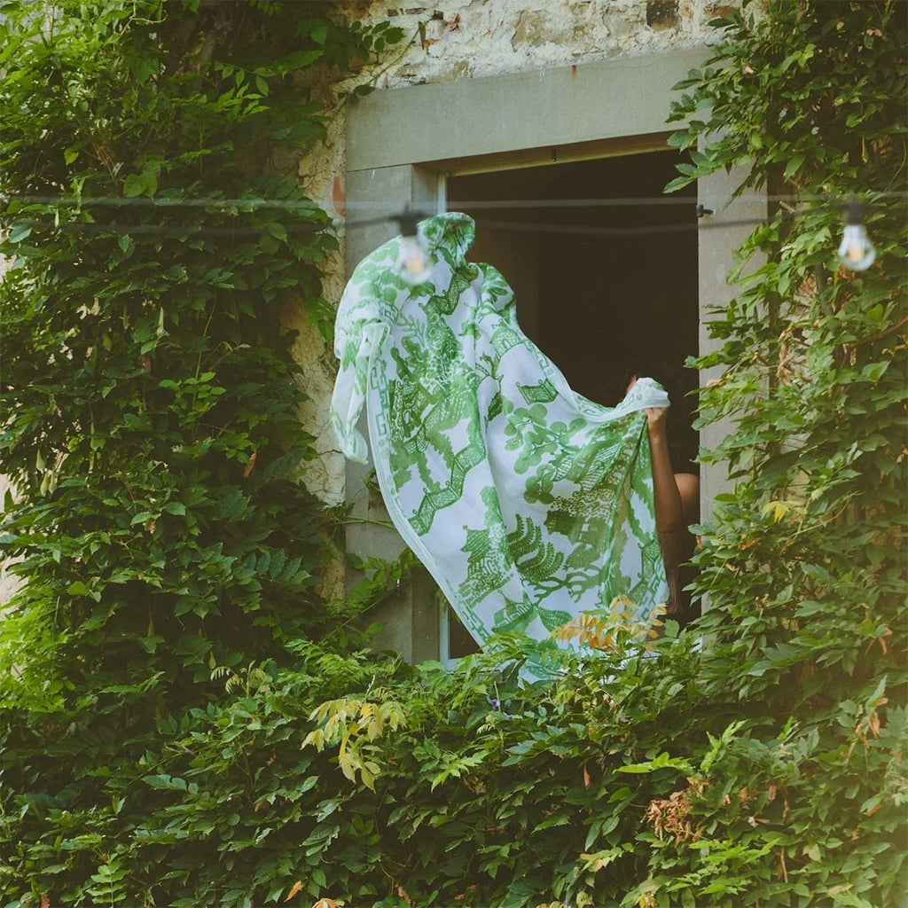 Lifestyle shot of green patterned silk scarf on clothing line hanging out of window surrounded by greenery and leaves. 