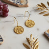 Gold earrings on a marble surface with grapes and decorative Mediterranean elements.