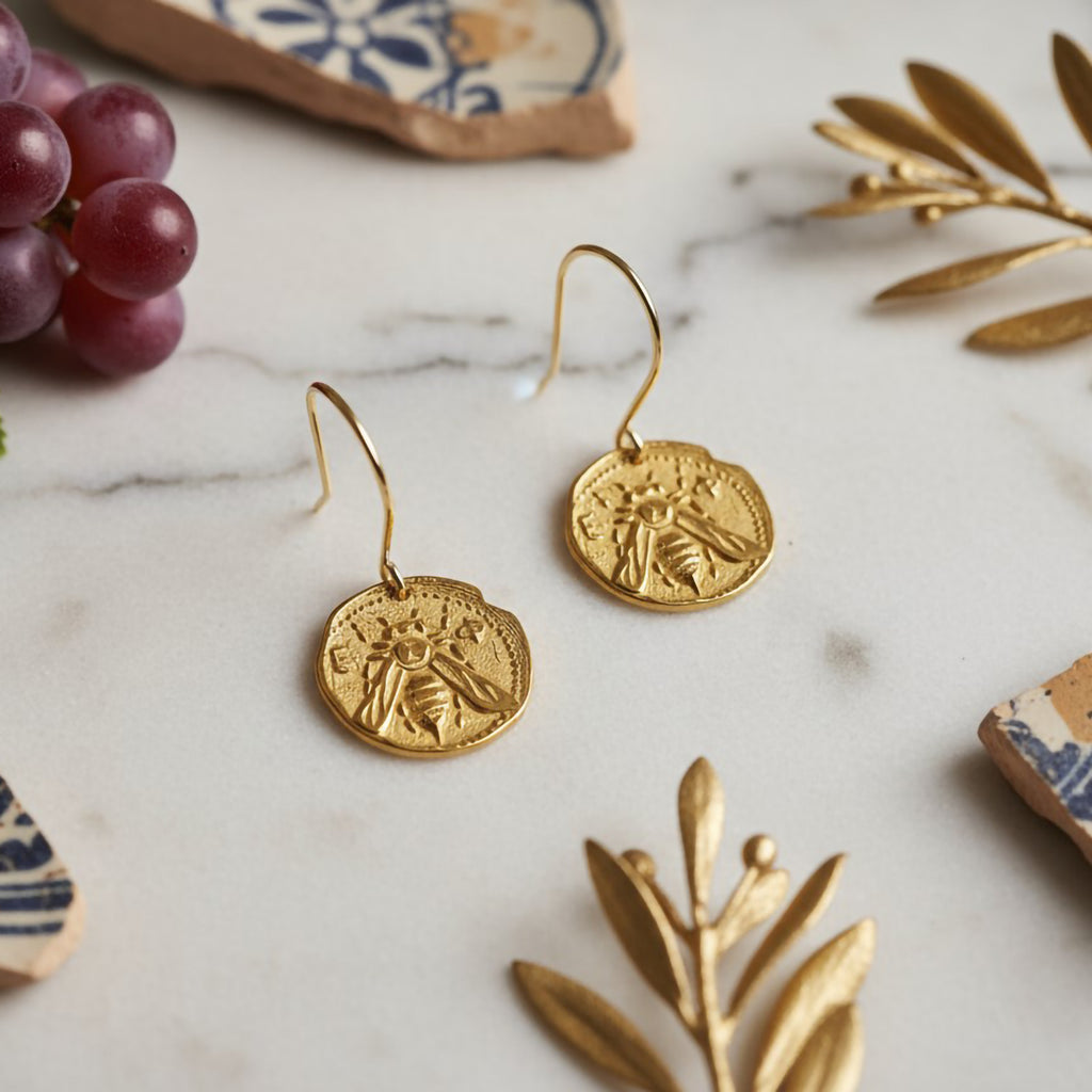 Gold earrings on a marble surface with grapes and decorative Mediterranean elements.