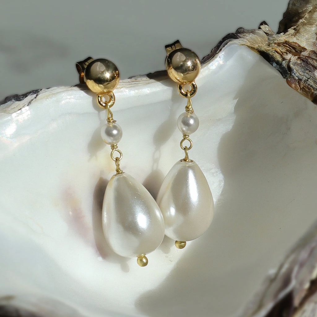 Close up of gold and pearl stud drop earrings resting on pearl coloured inside of oyster shell against a blurred background.