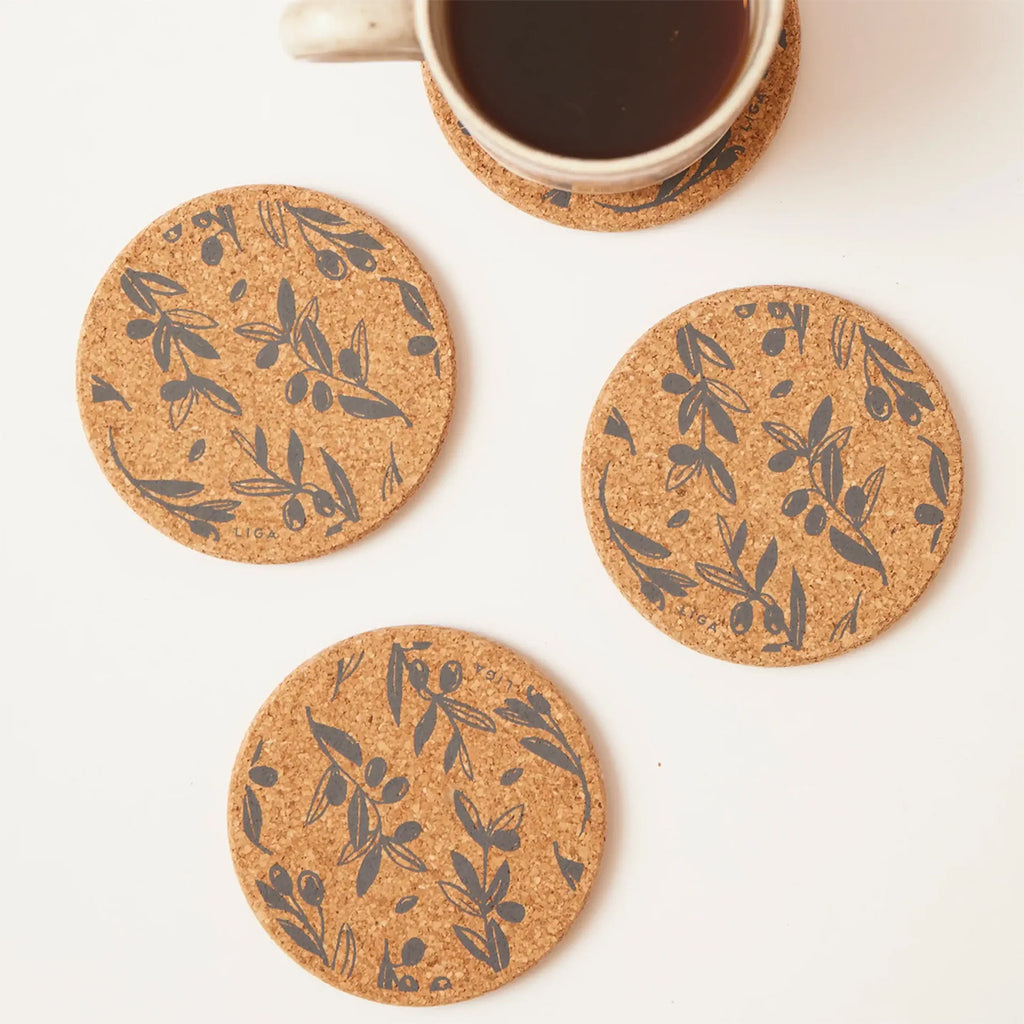 Four round cork coasters with a dark olive branch pattern are arranged on a white surface, with a white mug partially visible at the top on one of the coasters.