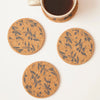 Four round cork coasters with a dark olive branch pattern are arranged on a white surface, with a white mug partially visible at the top on one of the coasters.