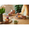A lifestyle scene of a couple sharing a breakfast at a wooden table, using matching round coasters and placemats with a grey olive leaf pattern. The table is decorated with croissants, a small jar of jam, a green mug, and a vase of pink tulips.