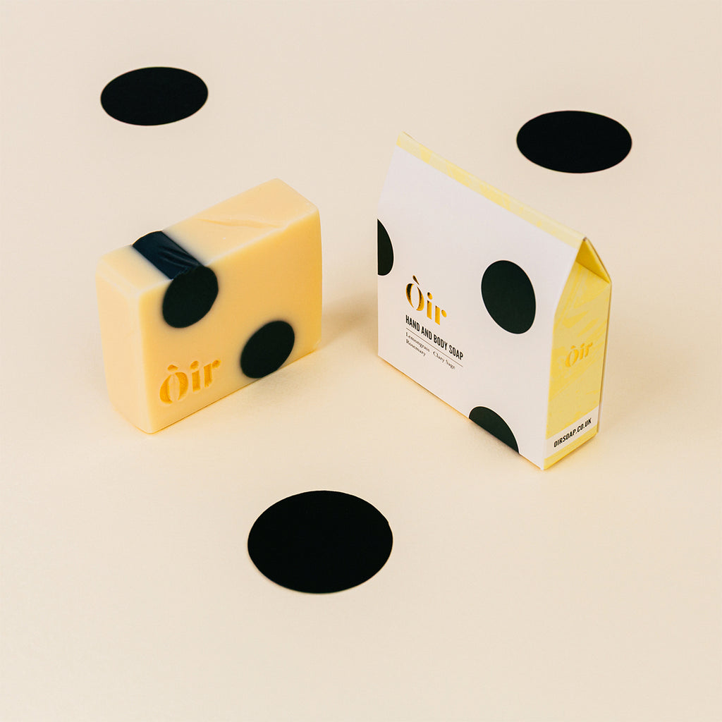 Yellow soap bar with black circular designs next to its packaging on a beige background