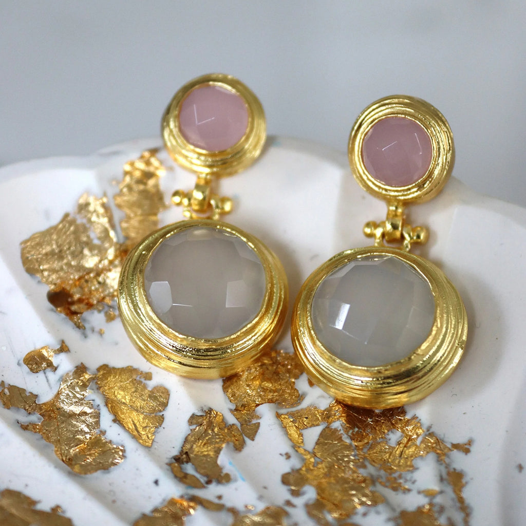 Close up of gold gemstone earrings with pink and white stones resting on marbled white surface with gold leaf flakes on a blurred backorund.