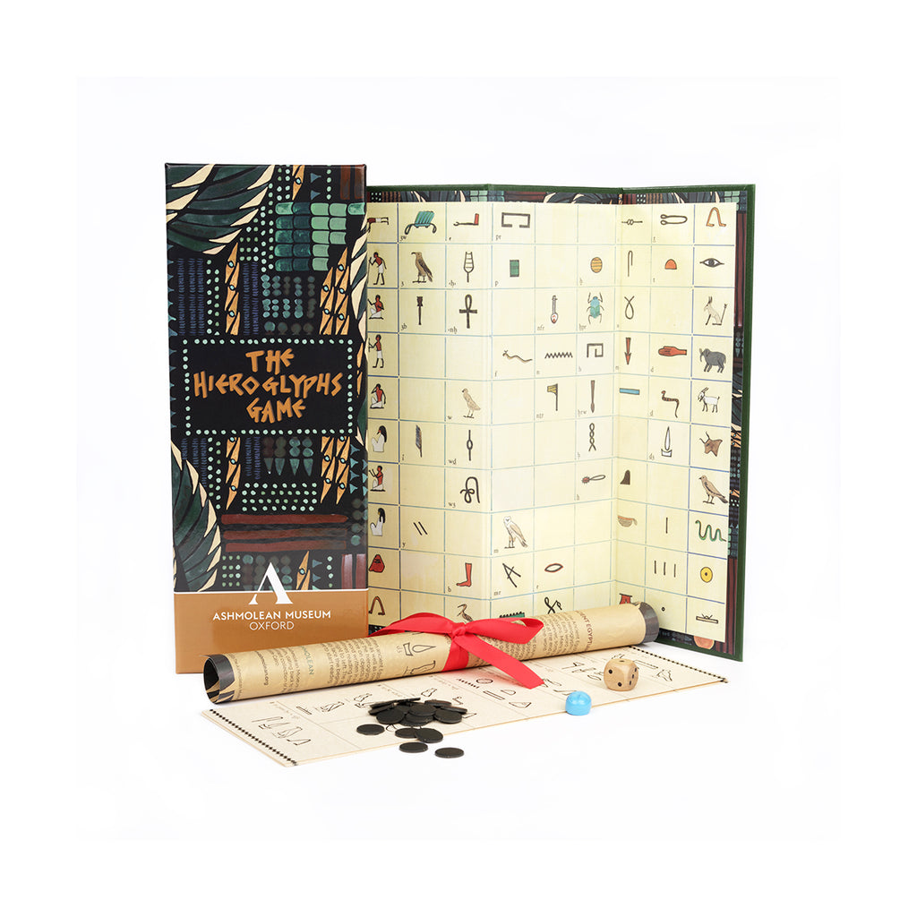 The Hieroglyphs Game set from the Ashmolean Museum, featuring a decorative box, an open game board with many engraved symbols, a rolled parchment scroll tied with a red ribbon, coins, and dice on a white background.