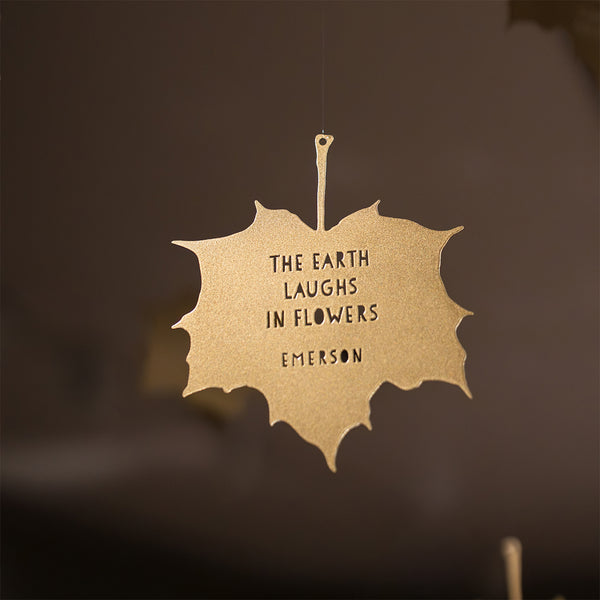 Close up focus of shiny gold metal maple leaf decoration hanging from wire against a blurred brown background