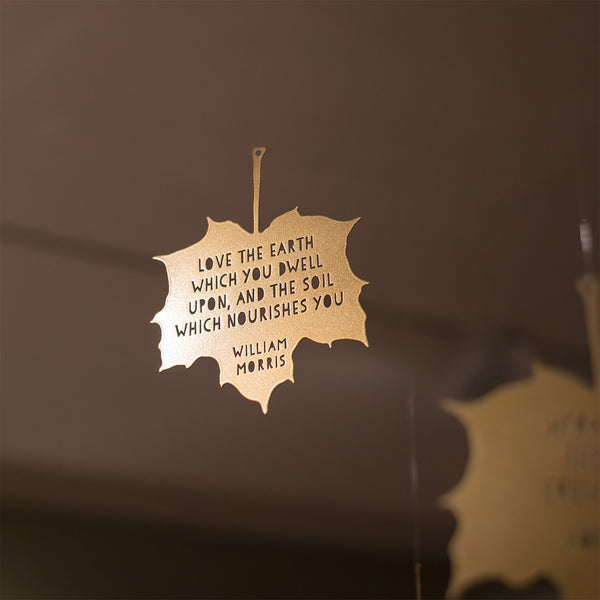 Close up focus of shiny gold metal maple leaf decoration hanging from wire against a blurred brown background
