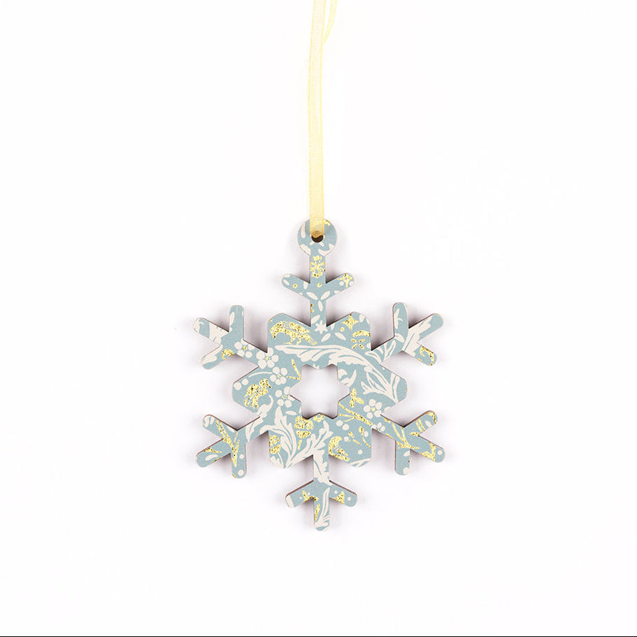 A decorative snowflake ornament with a light blue and white floral pattern and small gold accents, hanging from a pale gold ribbon on a white background.