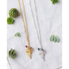 Two fern leaf necklaces, one in gold and one in silver with gemstone beads on white cotton cloth background surrounded by green succulent plants.