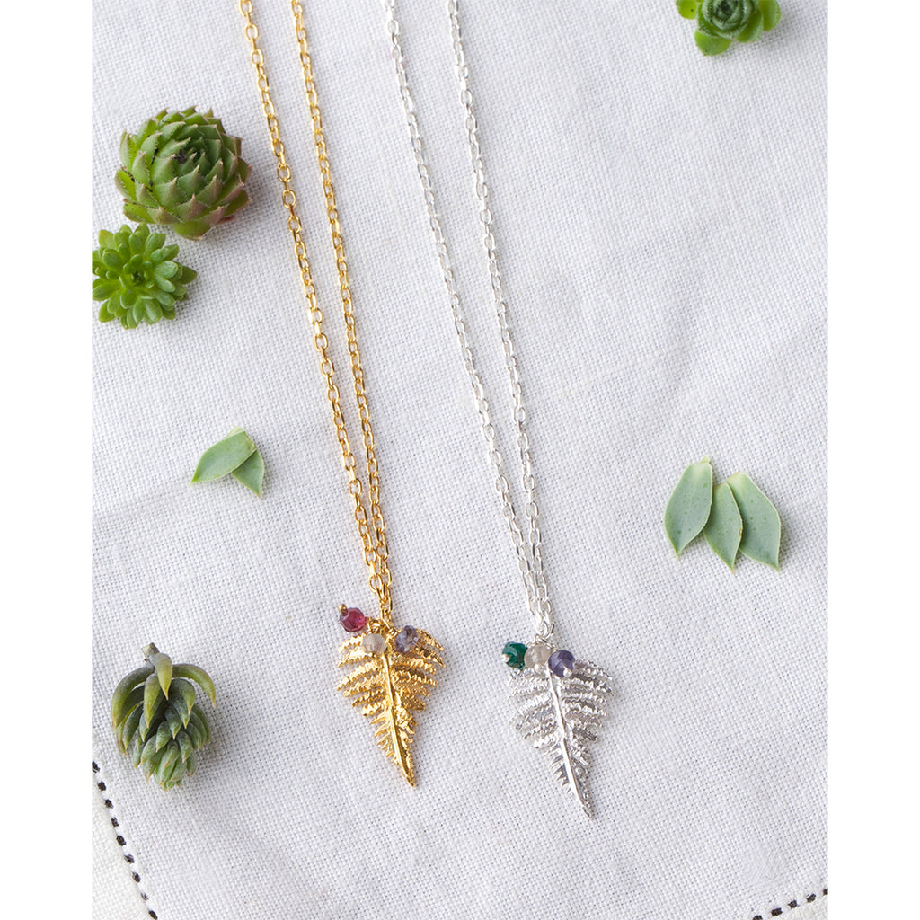Two fern leaf necklaces, one in gold and one in silver with gemstone beads on white cotton cloth background surrounded by green succulent plants.