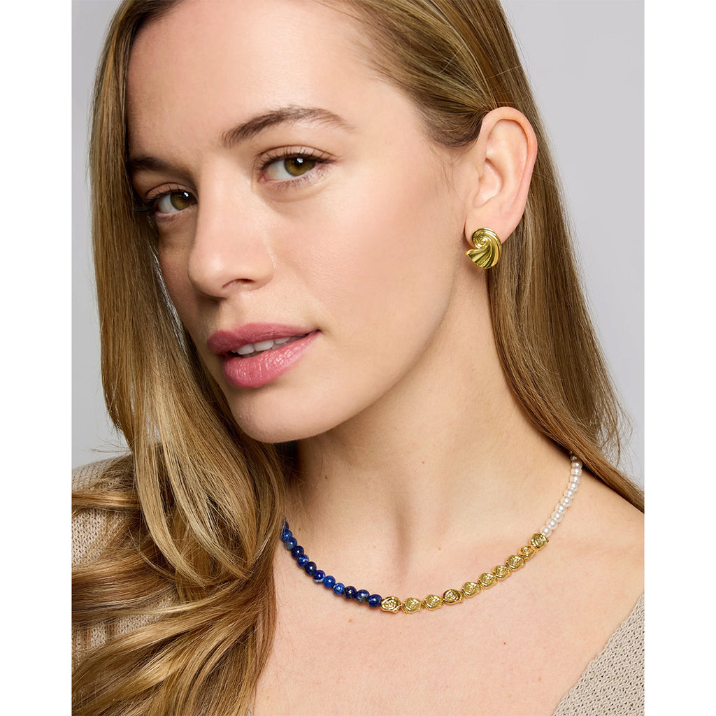 A close-up of a woman wearing a delicate necklace with blue, gold, and white beads, along with a matching gold earring, set against a neutral gray background.