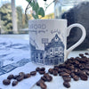 White china mug with design of blue Oxford buildings and University architecture and 'SILENCE PLEASE' sign, on top of table next to coffee beans.