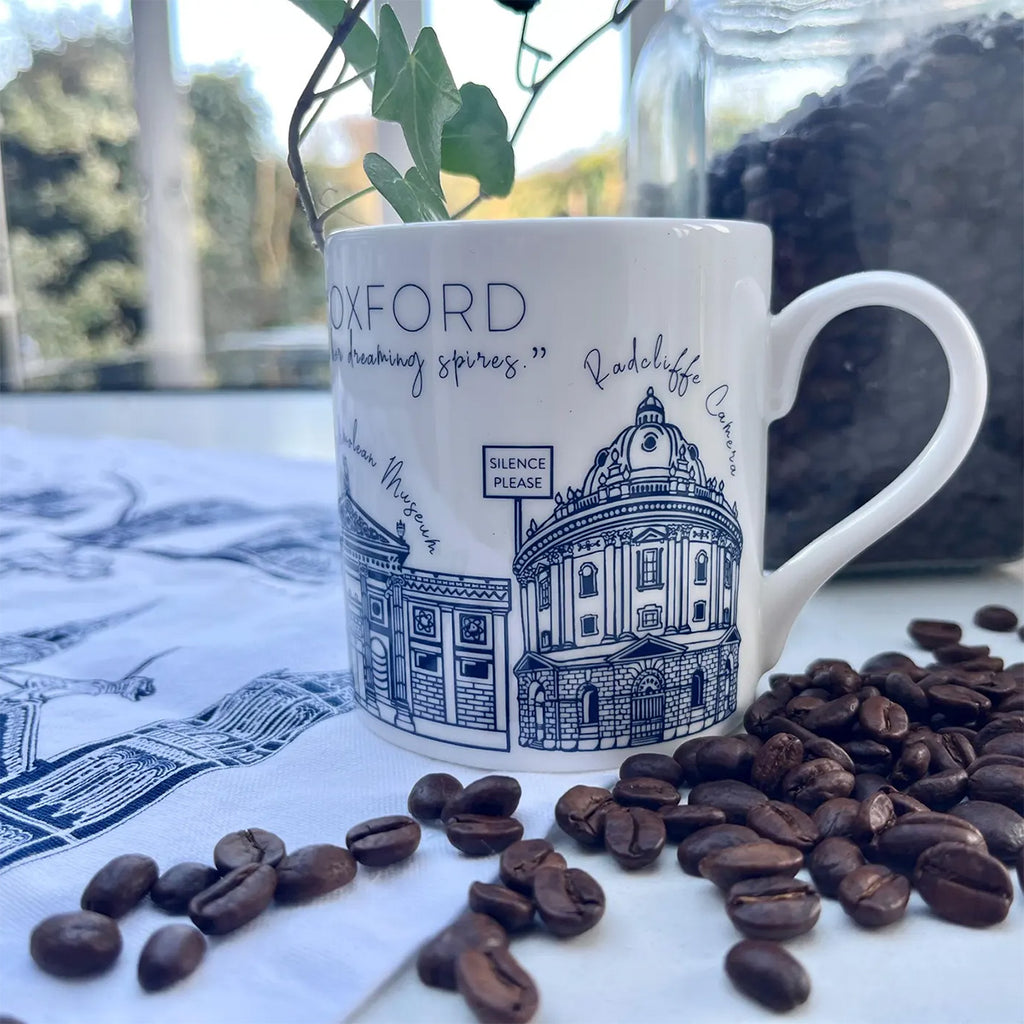 White china mug with design of blue Oxford buildings and University architecture and 'SILENCE PLEASE' sign, on top of table next to coffee beans.