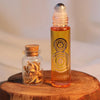 A glass roller bottle filled with amber-orange liquid stands on a wooden surface beside a small clear glass vial with a cork stopper containing dried curled botanical pieces. The main bottle has a silver cap and a printed gold target-like symbol, with the brand name ALASHIYA visible near the bottom.