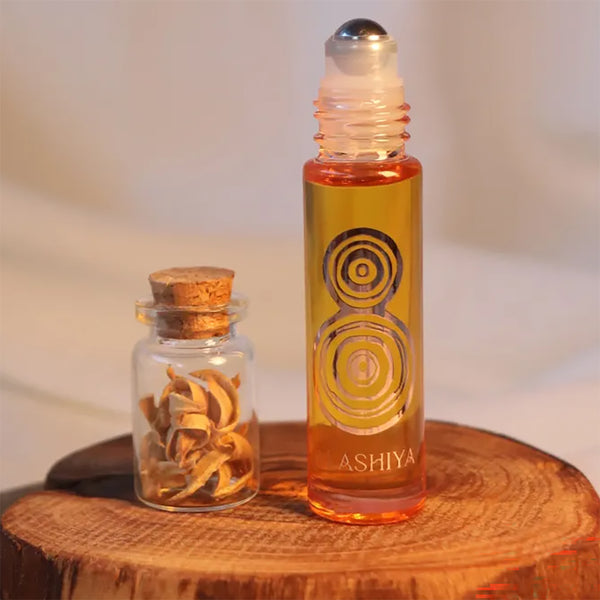 A glass roller bottle filled with amber-orange liquid stands on a wooden surface beside a small clear glass vial with a cork stopper containing dried curled botanical pieces. The main bottle has a silver cap and a printed gold target-like symbol, with the brand name ALASHIYA visible near the bottom.