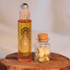 A small glass roller bottle filled with amber-orange perfume liquid stands beside a small glass jar with a cork stopper containing dried flower buds, both placed on a round wooden slice against a soft light background. The roller bottle has a black circular target-style graphic and the word 'ALASHIYA' near the bottom.