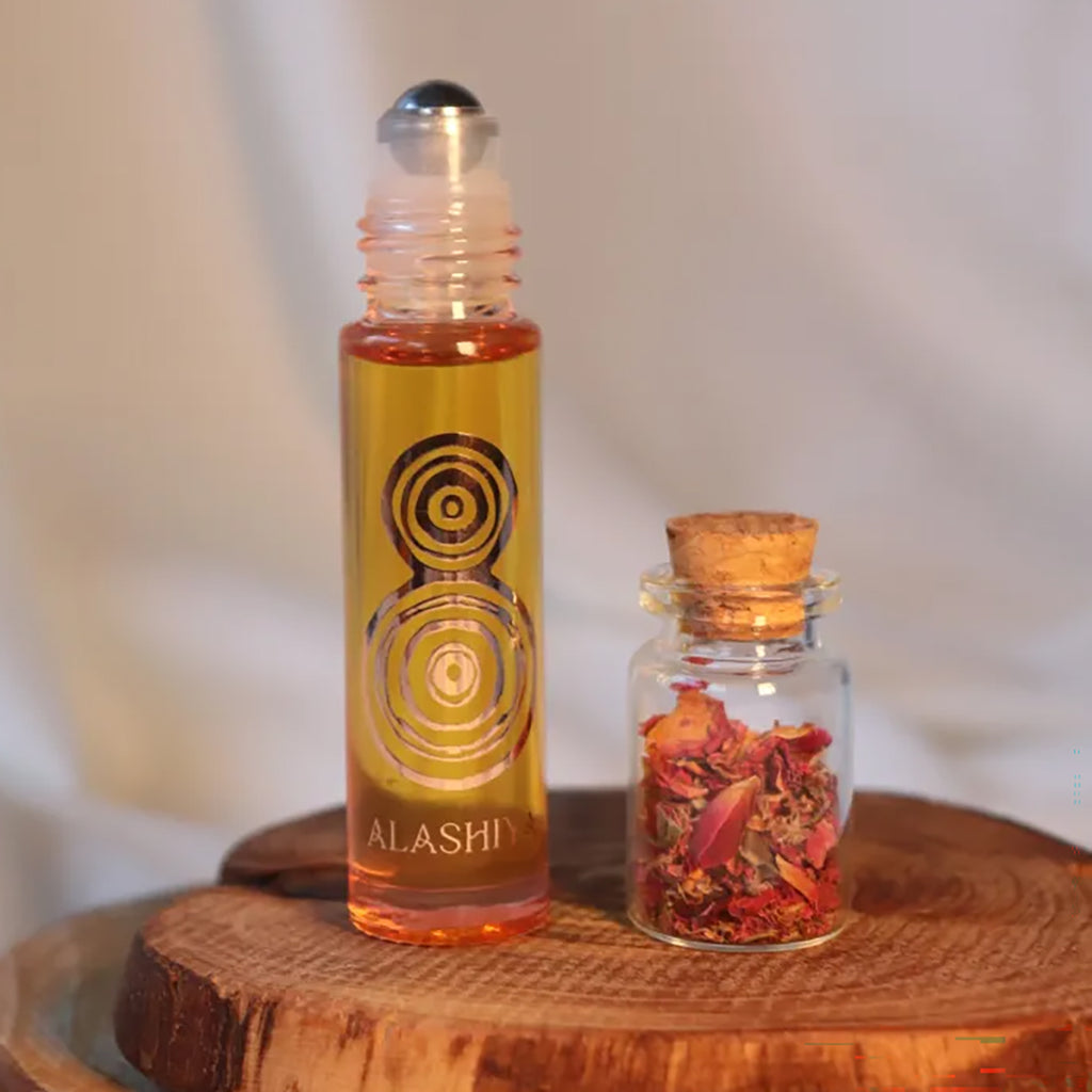 A roll-on bottle filled with amber-yellow perfume oil stands beside a small glass jar with a cork stopper and dried red botanical pieces inside, both placed on a round wooden slice against a soft light background. The roll-on bottle has a black circular graphic and the word 'ALASHIYA' near the bottom.