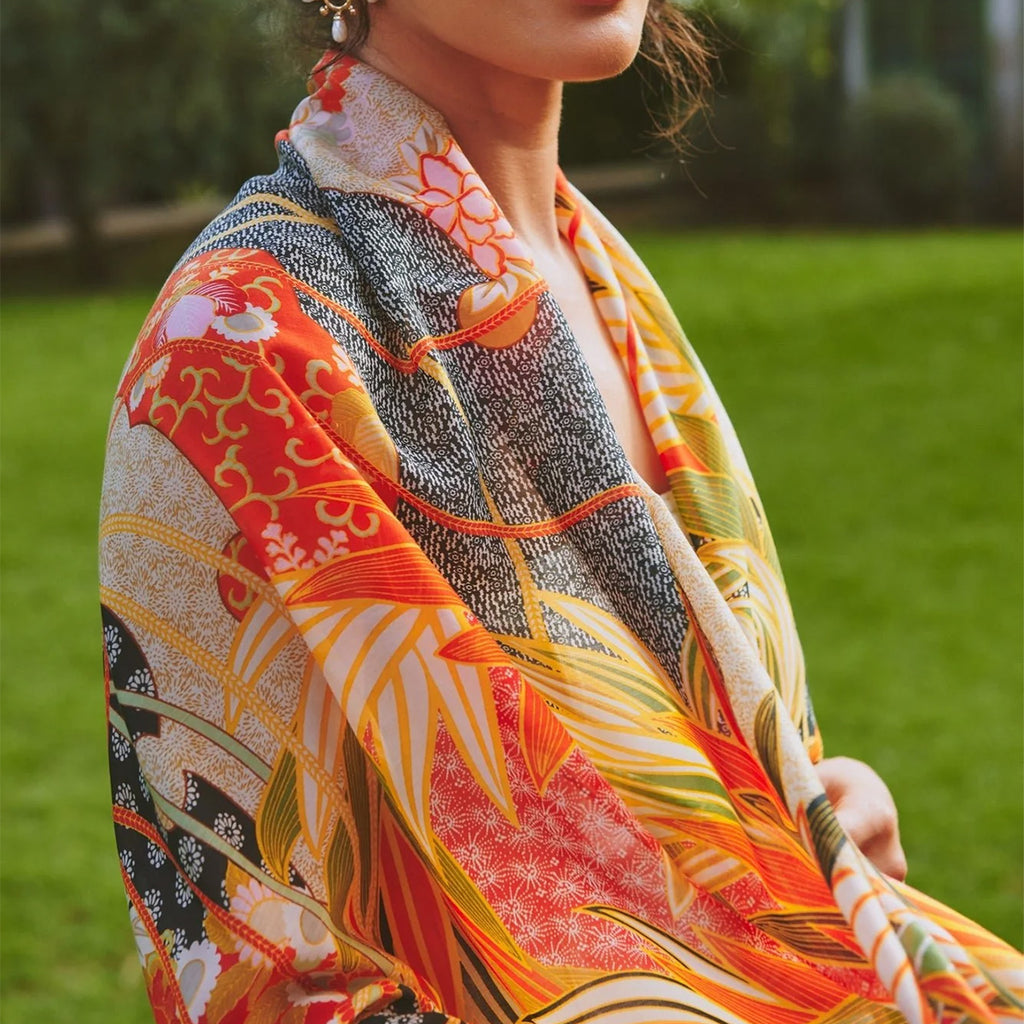 Model wearing floral patterned orange scarf over shoulders in foreground with grass and trees in background.