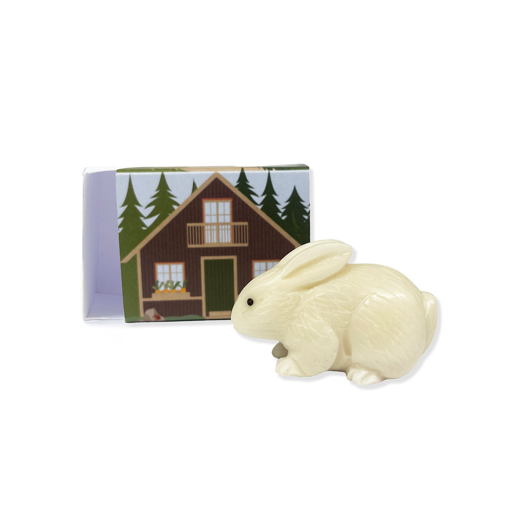 White carved miniature rabbit figurine in front of matchbox with woodland cabin artwork on a white background.