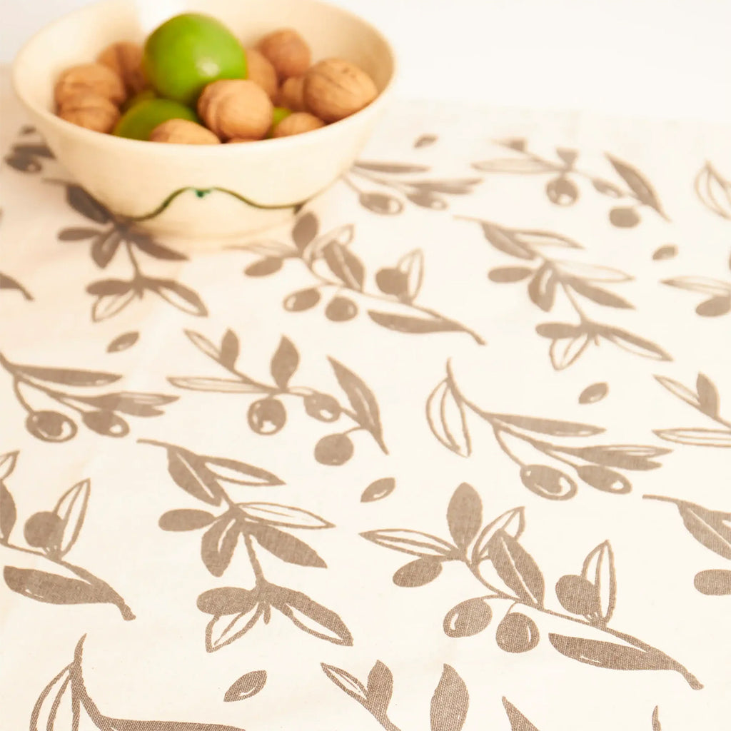 An off-white tablecloth with a repeating brown olive branch pattern, styled with a small bowl of walnuts and green apples in the upper left corner.