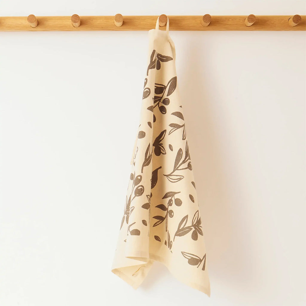 A cream-colored kitchen towel with a dark grey olive branch pattern, hanging from a wooden peg on a light wall.