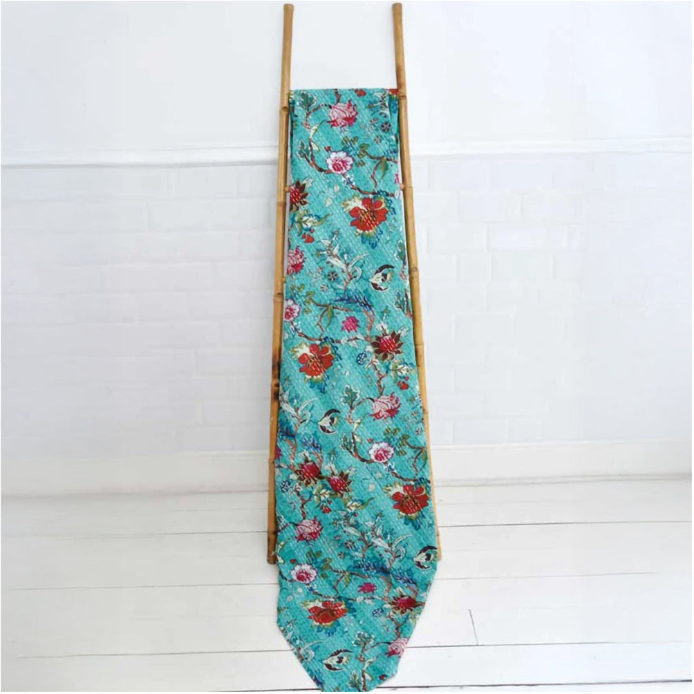 Floral-patterned teal cotton stitched throw blanket draped over wooden ladder against a white background