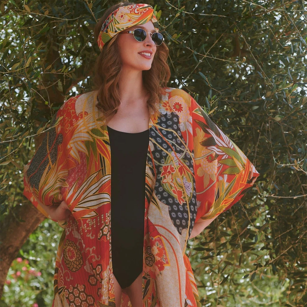 Summer lifestyle photo of female model wearing sunglasses and scarlet fans throwover over black swimsuit against olive trees background.