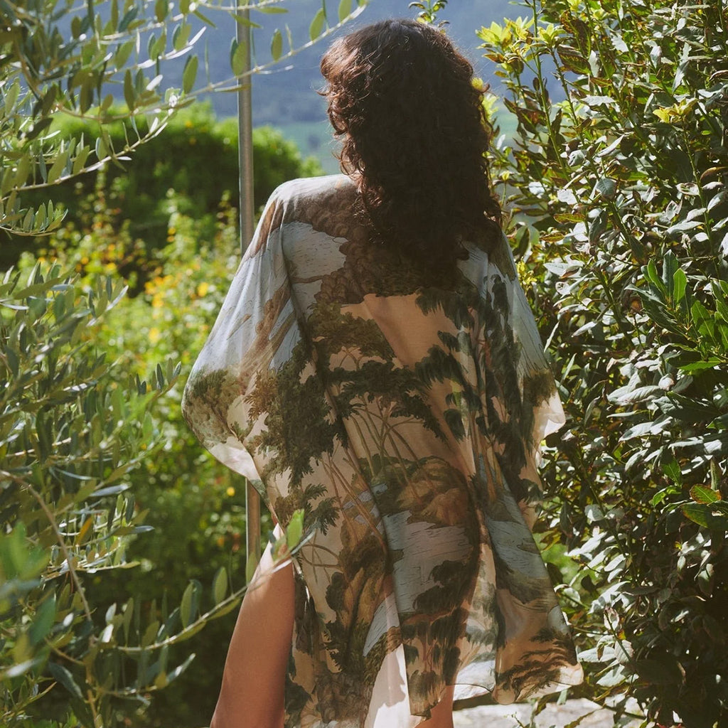 Model turned around to show back of rivers and trees pattern throwover standing amidst poolside summer olive trees and flowers.