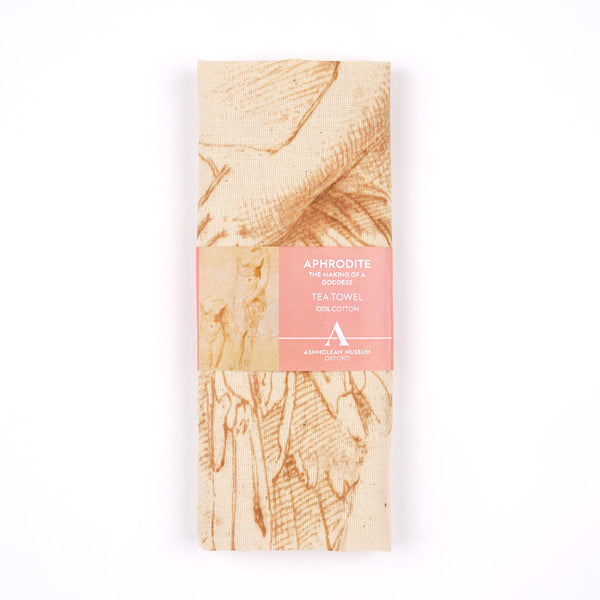 A folded cotton tea towel with a beige linen-like texture and brown sketch-style illustration, wrapped in a pink paper band labeled 'APHRODITE THE GARDEN OF GODS TEA TOWEL 100% COTTON' on a white background.