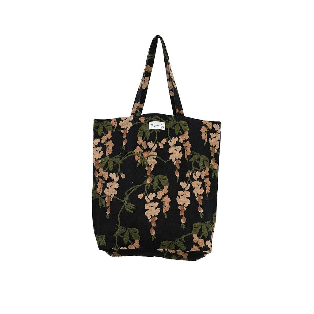 A black canvas tote bag with a wisteria floral print in shades of beige and olive green, featuring dual shoulder straps and a small white brand label at the top center.