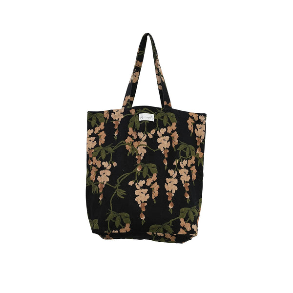 A black canvas tote bag with a wisteria floral print in shades of beige and olive green, featuring dual shoulder straps and a small white brand label at the top center.