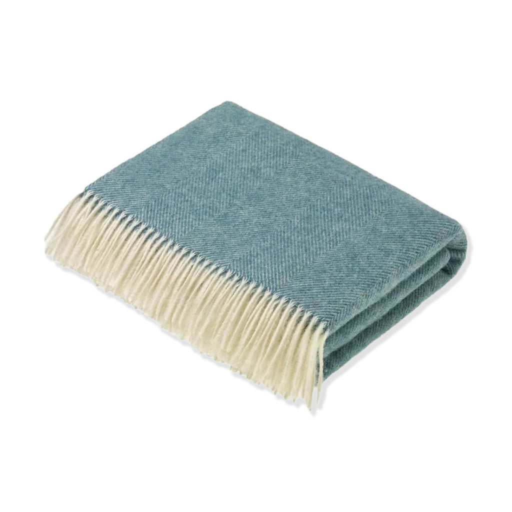 Folded wool throw in green/blue herringbone pattern with white tasselled edges on a white background.