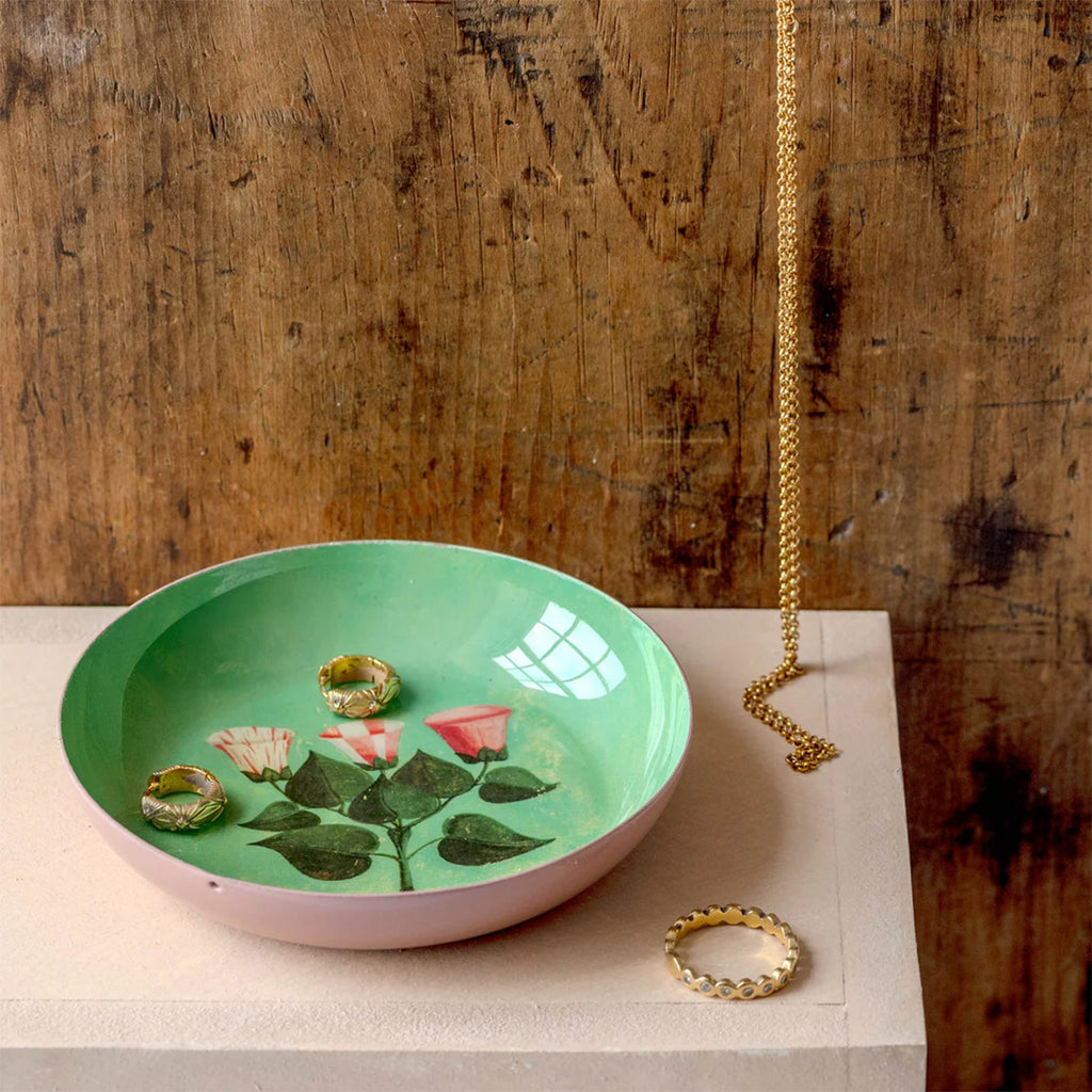 Lifestyle photo of round enamel tray being used to hold gold rings and necklace jewellery on pink table with wood background.